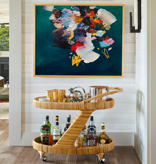 Made Goods Lulu wrapped rattan bar cart  styled with glasses, bottles, ice bucket, etc. in a room with shiplap paneling and an abstract painting.