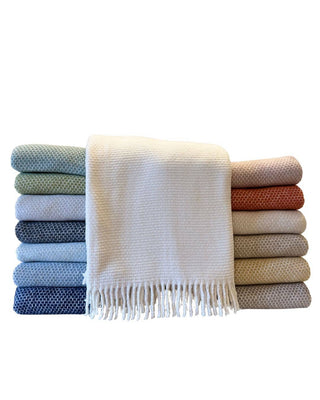 A stack of folded woven throw blankets in multiple colors, with a cream-colored fringed throw draped across the center, shown on a plain white background.