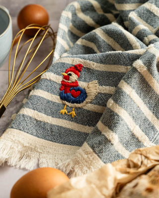 Striped towel with embroidered chicken design on a kitchen counter with eggs and a whisk.