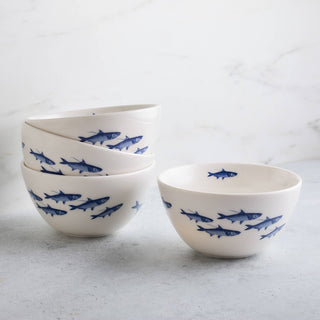A set of white ceramic bowls decorated with blue fish illustrations, with three stacked on the left and one standing on the right, set against a light gray marble background.