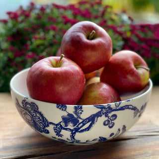 Caskata Arcadia Vegetable Bowl of red apples with a blue floral pattern on a wooden surface with blurred flowers in the background