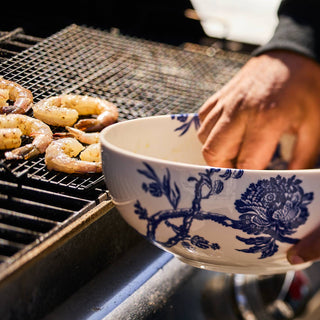 Person holding a Caskata Caskata Arcadia Vegetable bowl  blue and white floral-patterned bowl over a grill with shrimp cooking.