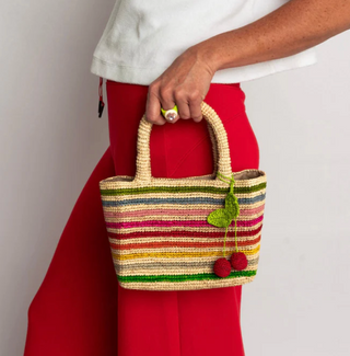Colorful striped handbag with cherries bag tag held by a person wearing red pants and a white top.