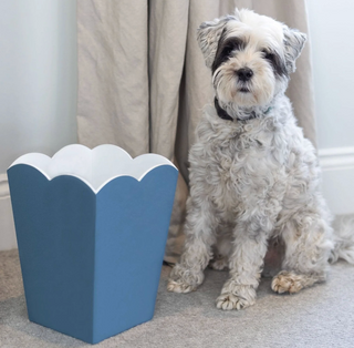 White dog sitting next to a blue decorative Addison Ross trash can with scalloped edges.
