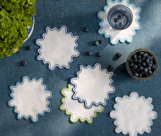 Decorative Matouk Linen Wave Cocktail Napkins with wavy edges on a textured surface with blueberries and green leaves.