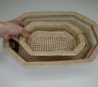 Three nested octagonal woven baskets made of natural beige rattan or seagrass, with a hand holding the smallest basket to show its depth, on a plain light gray background.