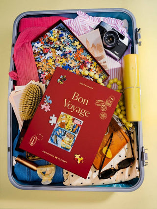 Piecework Puzzles Bon Voyage - Open suitcase with travel items including a 'Bon Voyage' book, puzzle, camera, and other small objects on a yellow background.