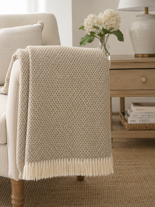A textured cream and beige woven throw blanket with fringe trim draped over the arm of a light-colored armchair in a softly styled living room setting.